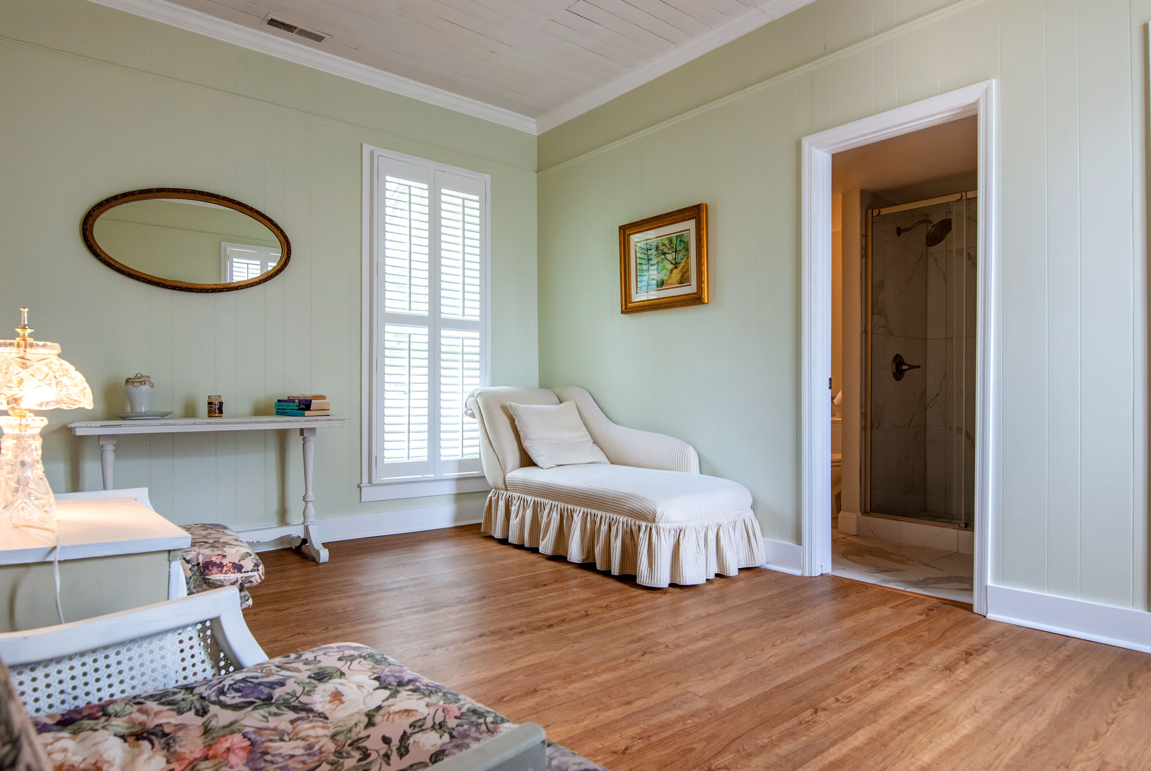 Una habitación con suelo de madera, un sofá blanco, un sillón estampado, una mesita con una lámpara, un cuadro en la pared y una puerta que da a una zona de ducha.