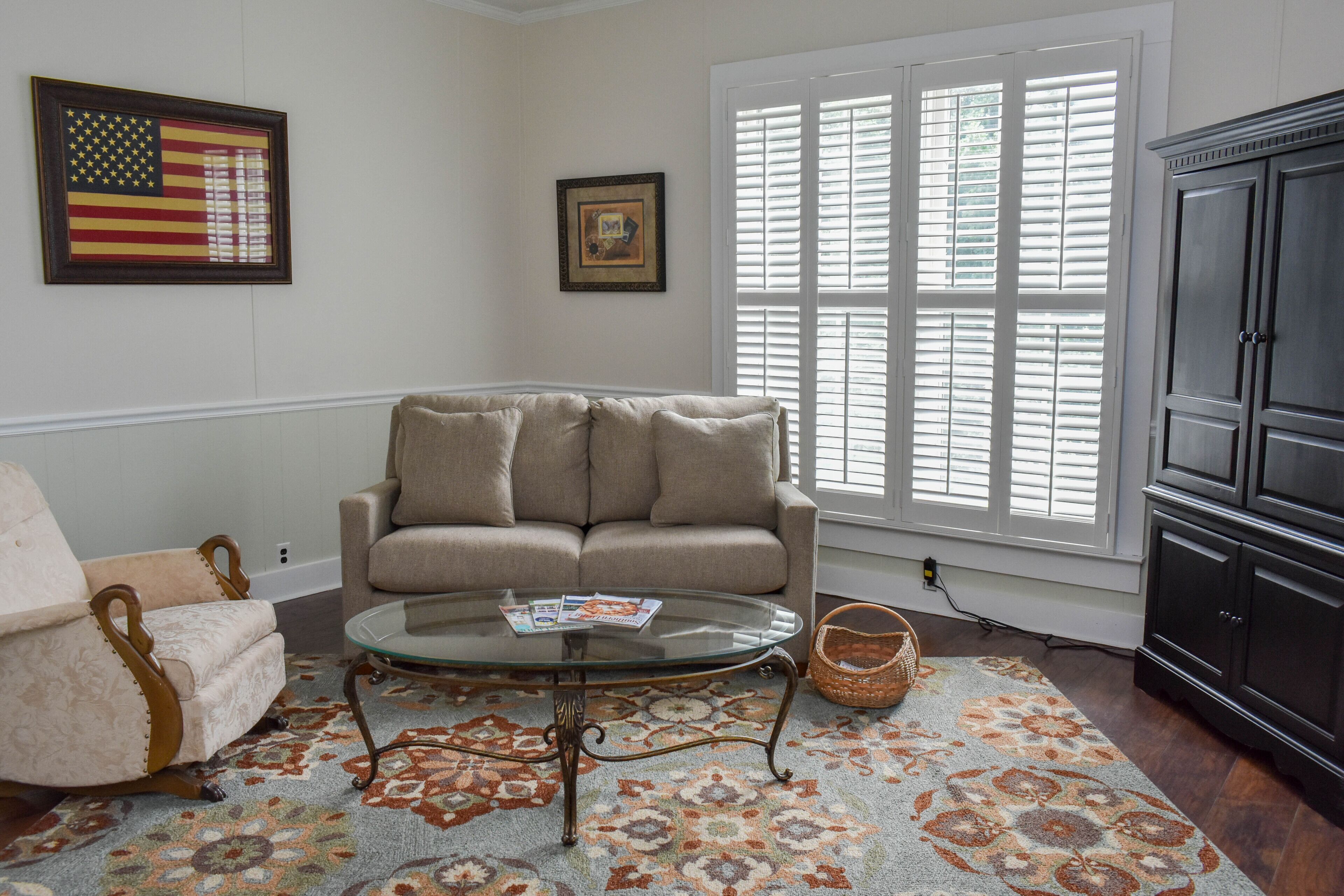 A living room with a beige sofa, a glass coffee table, a patterned rug, a dark wooden cabinet, and a window with white shutters.