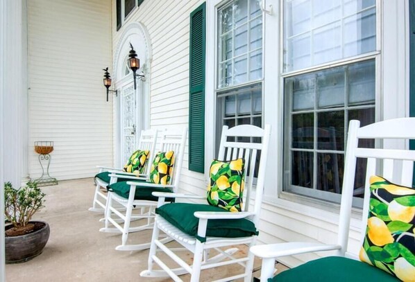 A porch with white rocking chairs and green cushions, a potted plant, and a window with shutters.