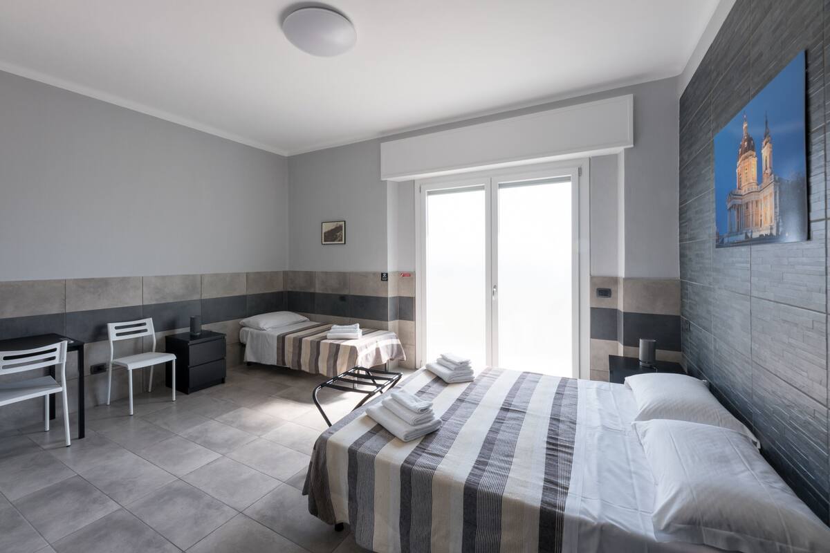 Una habitación de hotel con una cama, dos mesitas de noche, una silla, un escritorio y un ventanal grande.