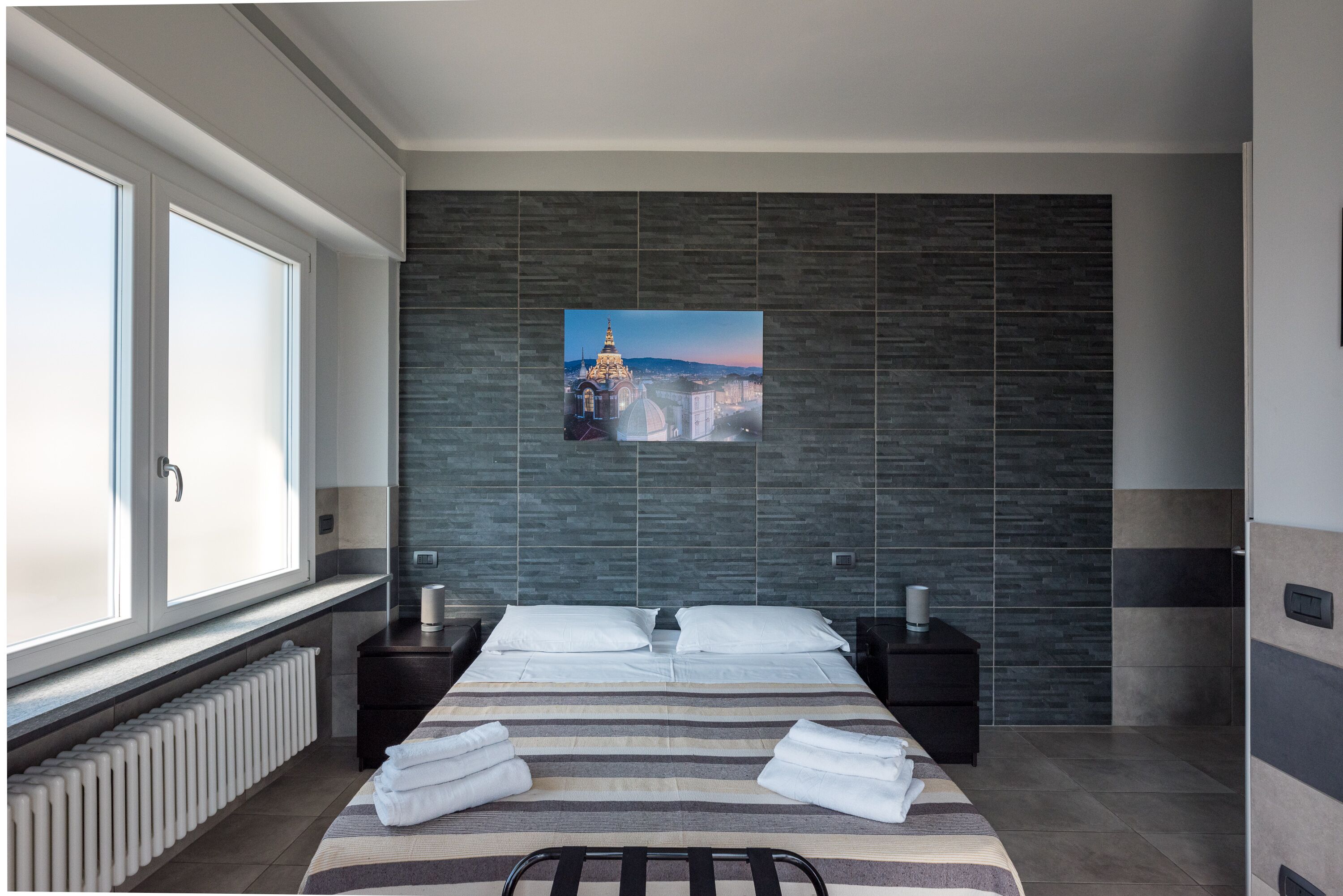 A modern hotel room with a large bed, a view of a cityscape, a dark tiled wall, and a window with blinds.