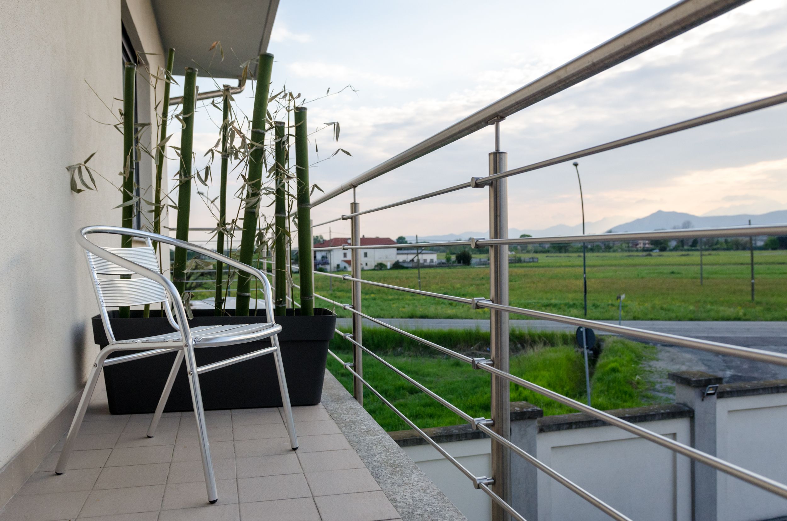 A balcony with a metal chair, a potted plant, and a view of a green field and distant mountains.
