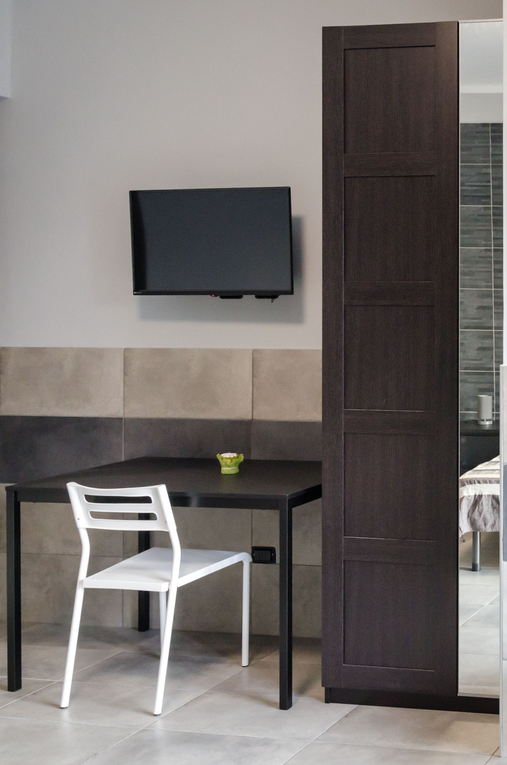A modern room with a dark wooden door, a white chair, a small table, and a flat-screen TV mounted on the wall.