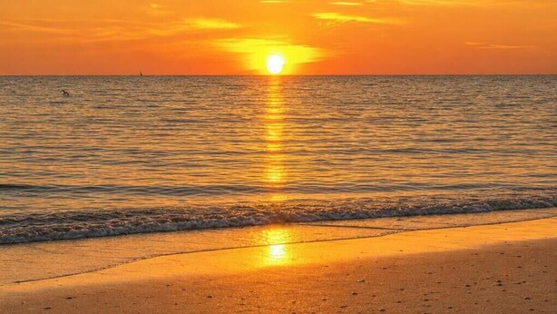 Een zonsondergang boven een strand, met rustig water dat de zon weerspiegelt.