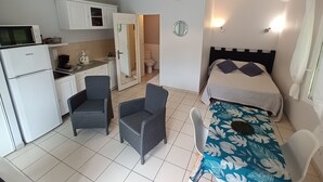 A compact room with a kitchenette, a bed with a floral bedspread, two chairs, a small table, and a ceiling light fixture.