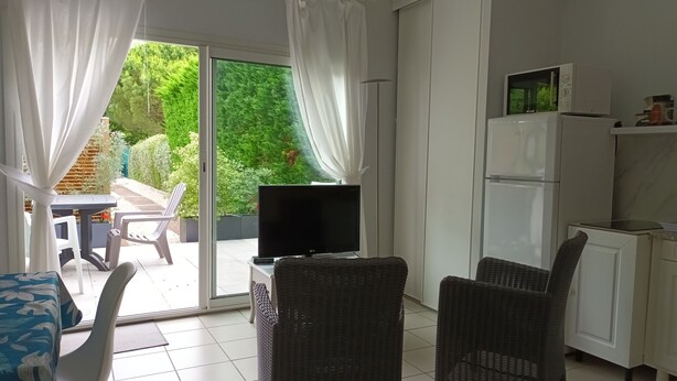 A room with a TV on a stand, wicker chairs, a dining table, and a kitchen area with a fridge and microwave.
