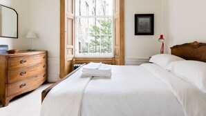 A neatly made bed with white linens, a wooden dresser with drawers, a red lamp, and a window with a view of buildings and trees. - Stylish City Centre Apartment (Edinburgh)