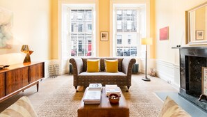 A living room with a sofa, a coffee table with books, a fireplace, and large windows with a city view. - Stylish City Centre Apartment (Edinburgh)