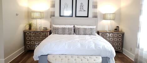 A bedroom with a large bed, two bedside tables with lamps, a bench, and two framed pictures on the wall.
