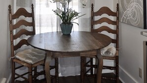 Un área de comedor con una mesa de madera redonda, cuatro sillas de madera, un jarrón con flores blancas y una ventana con cortinas de encaje.