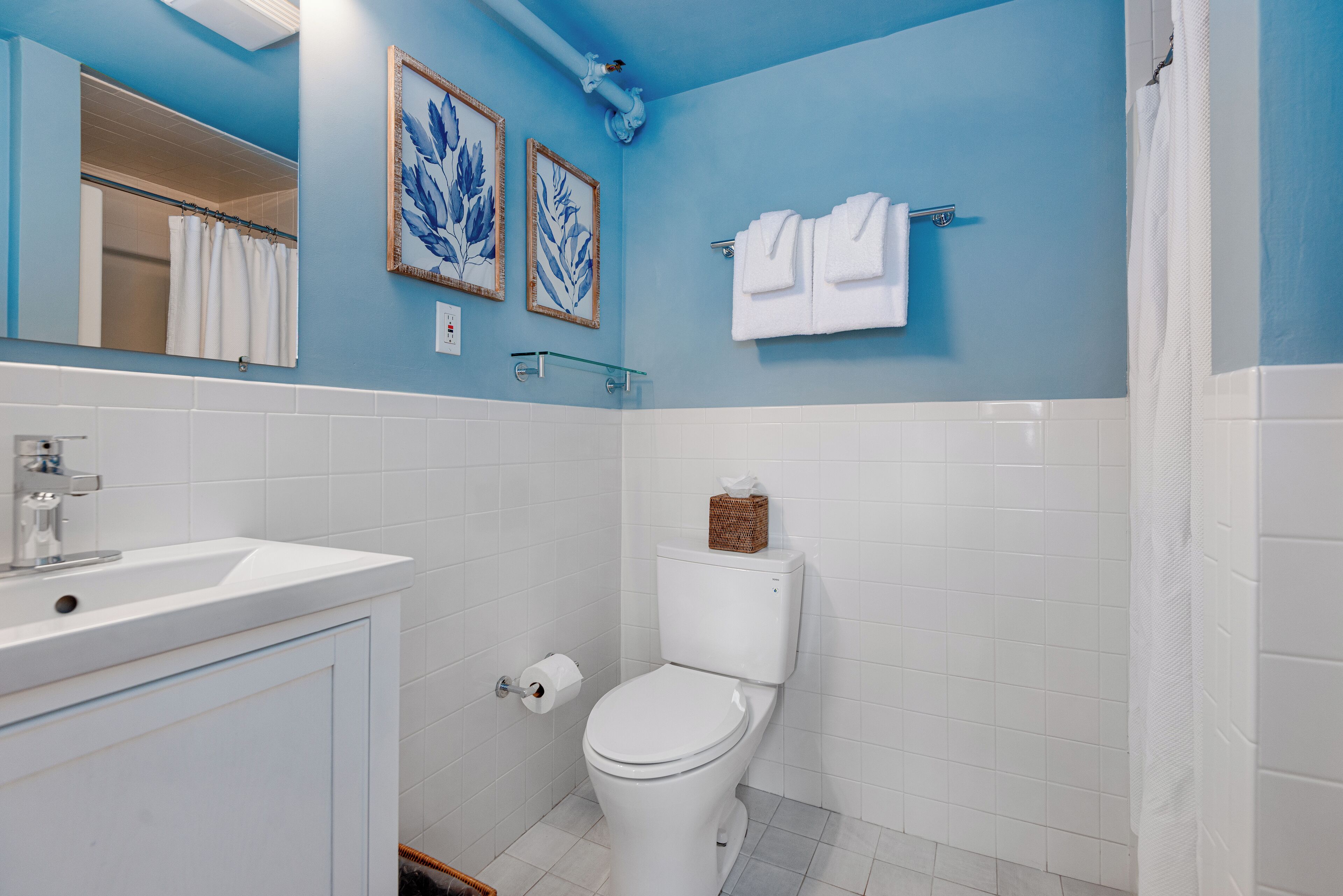 Un baño con lavamanos, inodoro y cortina de ducha blancos. En la pared hay cuadros con motivos florales.