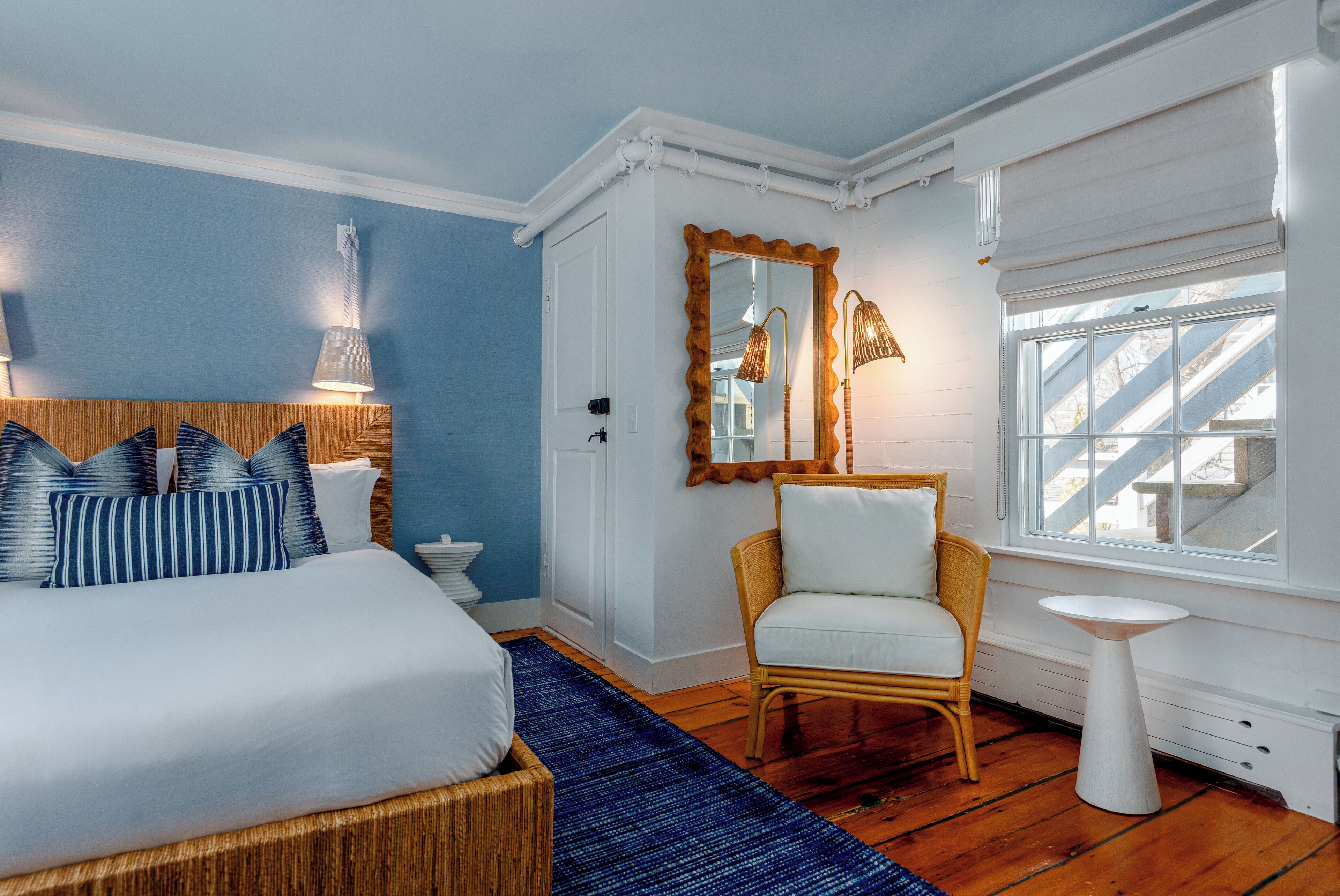 A bedroom with a bed, a chair, a small table, a mirror, and a window with a view of a staircase.