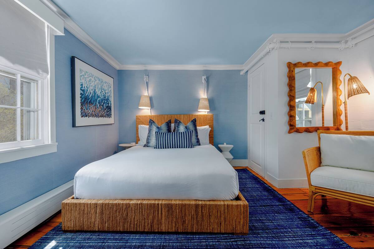 Quarto com uma cama grande, duas luminárias de cabeceira, um espelho com moldura decorativa e um banquinho de vime.