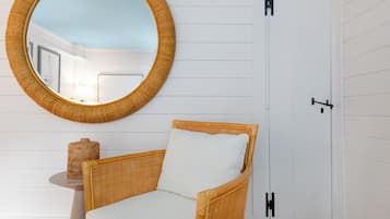 Una silla de mimbre con un cojín blanco, un espejo redondo y una mesita con un recipiente de mimbre.