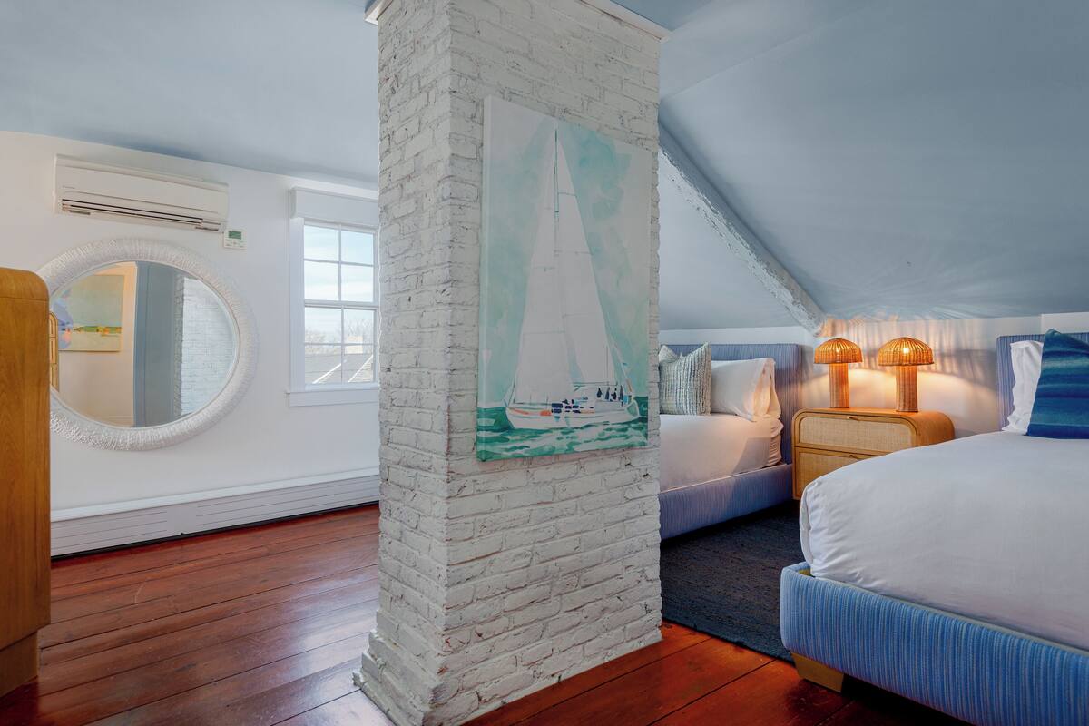 Um quarto com um pilar de tijolos brancos, um espelho redondo, uma pintura de um veleiro, uma cama com roupa de cama azul e uma mesa de cabeceira com duas luminárias.
