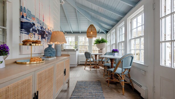 A bright kitchen with a high ceiling, wooden cabinets, a dining area with wicker chairs, and a large window providing natural light. - Anchor Inn (Nantucket)