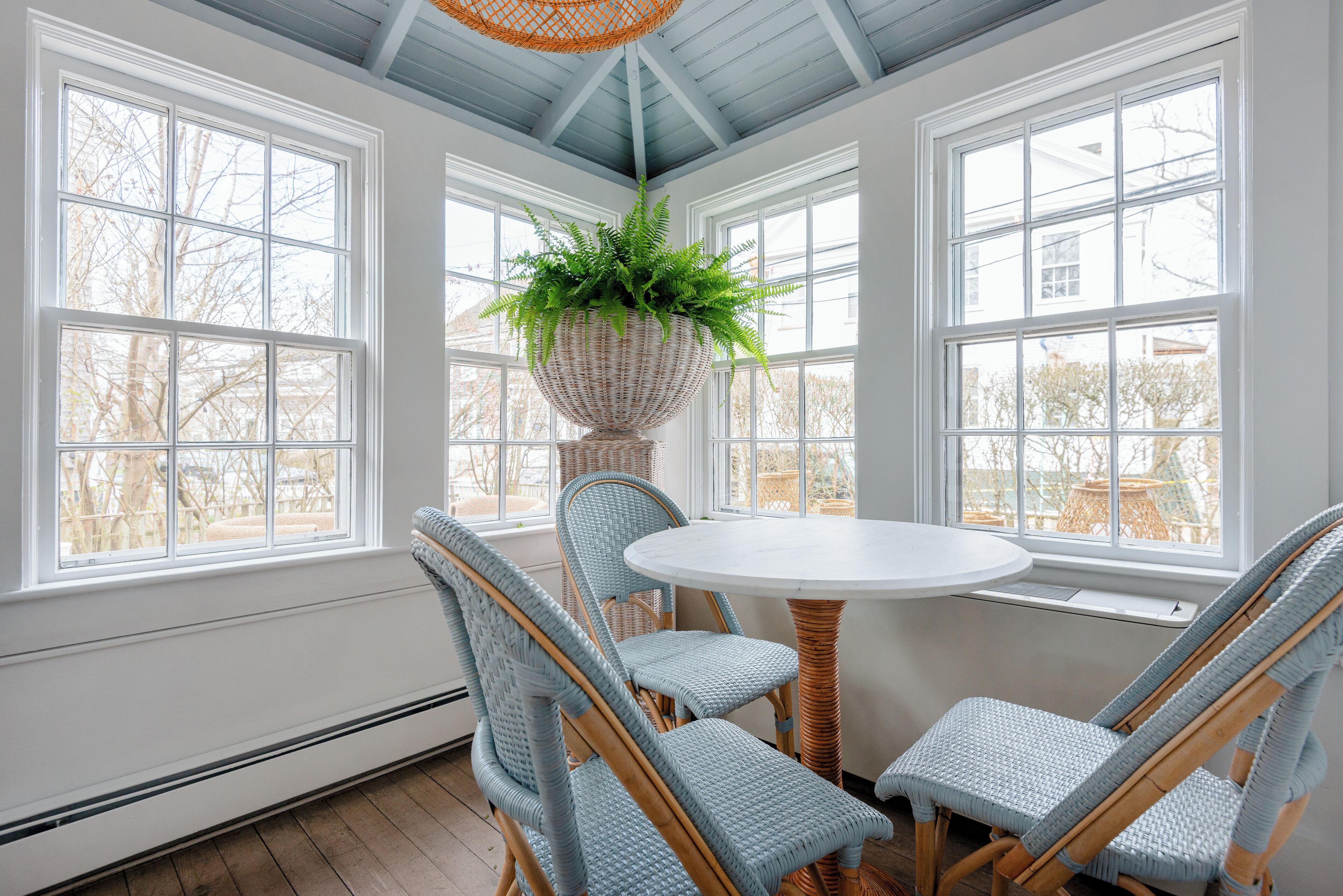 A sunroom with a round table and four wicker chairs, large windows, and a hanging plant.