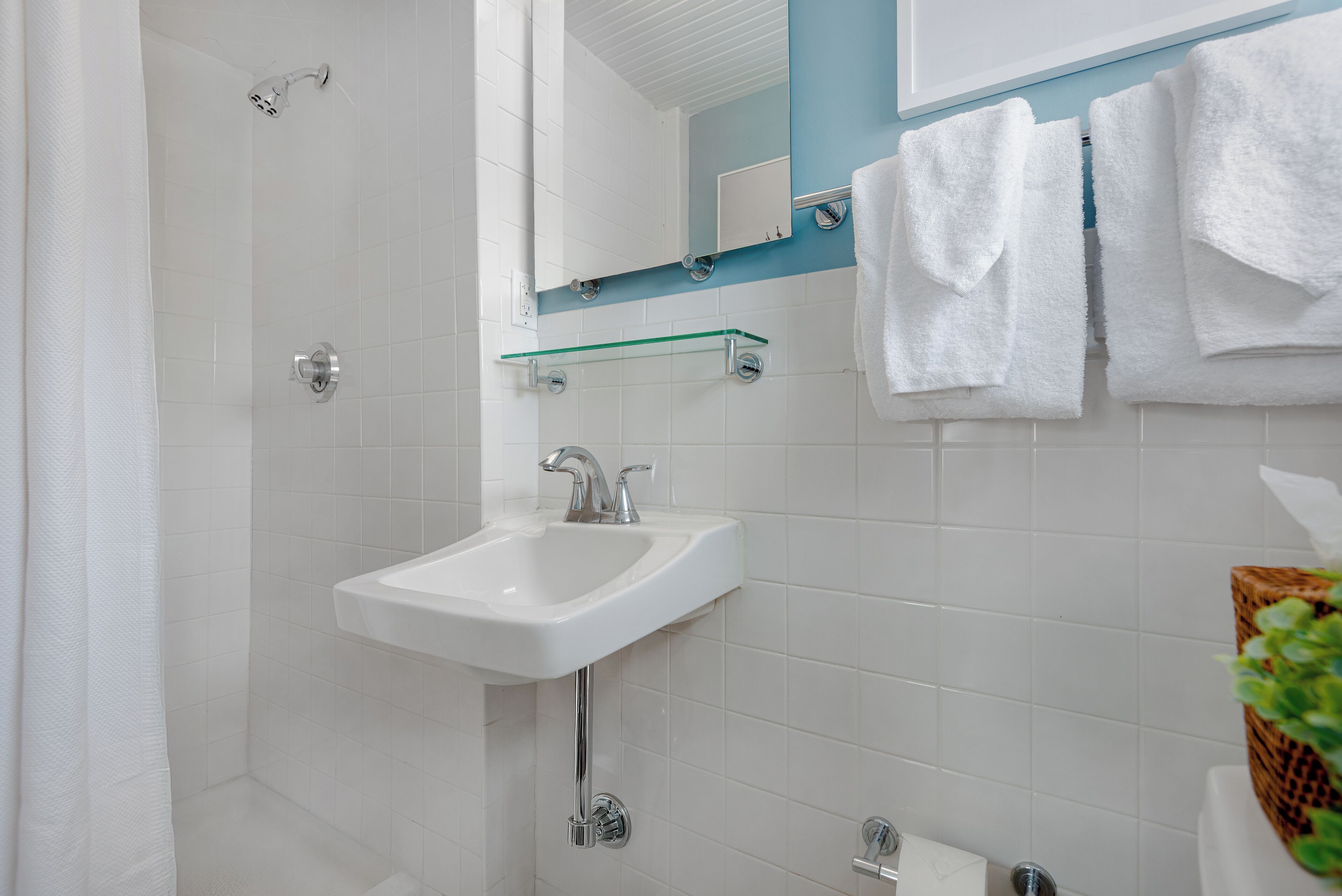 Un baño con un lavamanos blanco, un estante de vidrio, una ducha y toallas blancas colgadas en la pared.