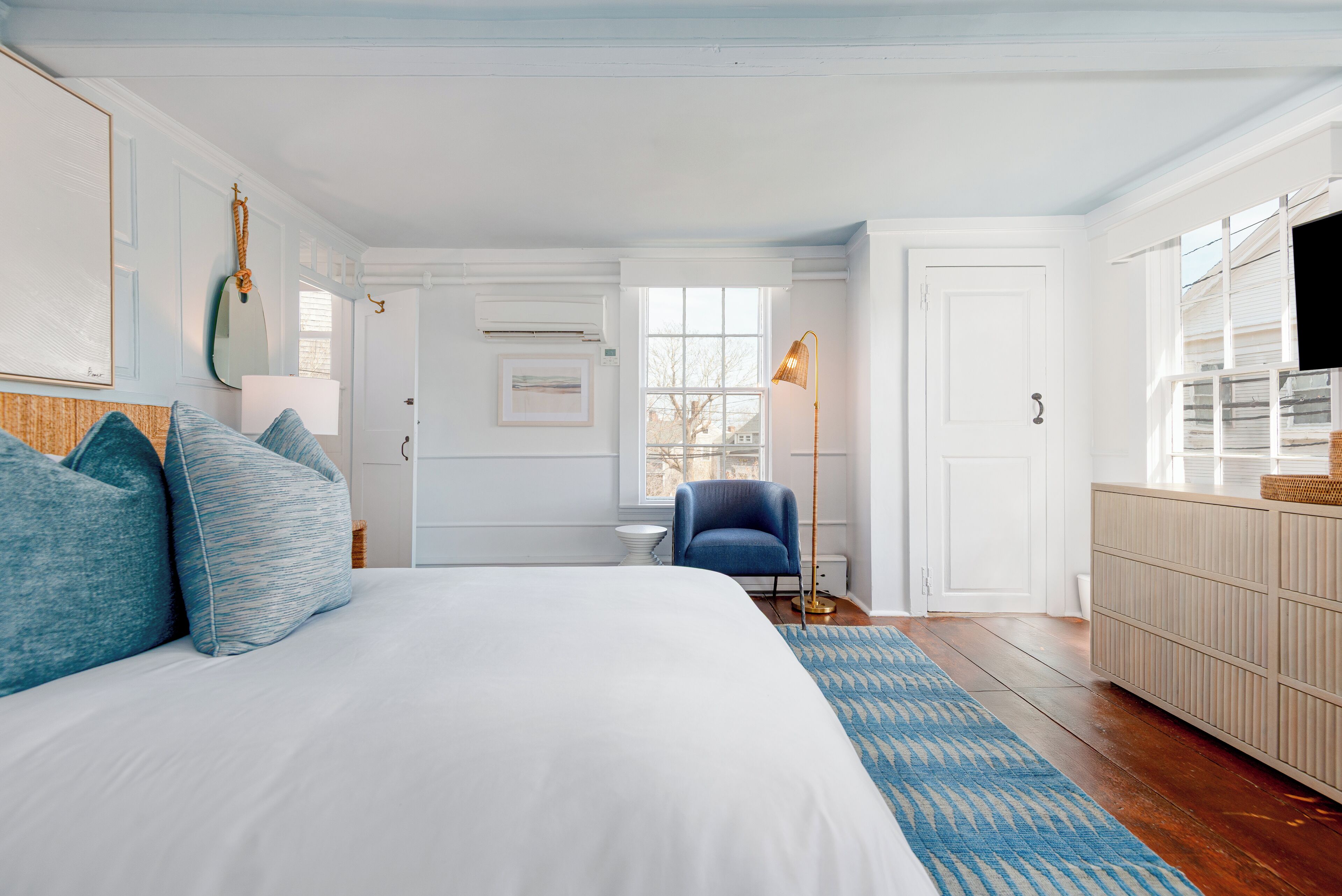 Un dormitorio con una cama grande, un sillón azul, una lámpara de pie y vistas al exterior a través de una ventana.