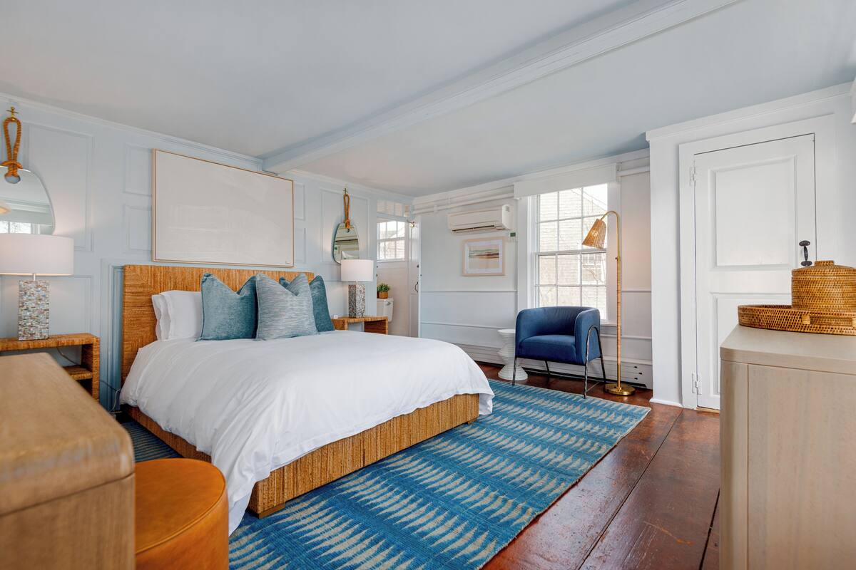 Quarto com uma cama grande, um tapete azul e branco estampado, uma cadeira azul e uma mesinha de cabeceira de madeira com uma luminária.