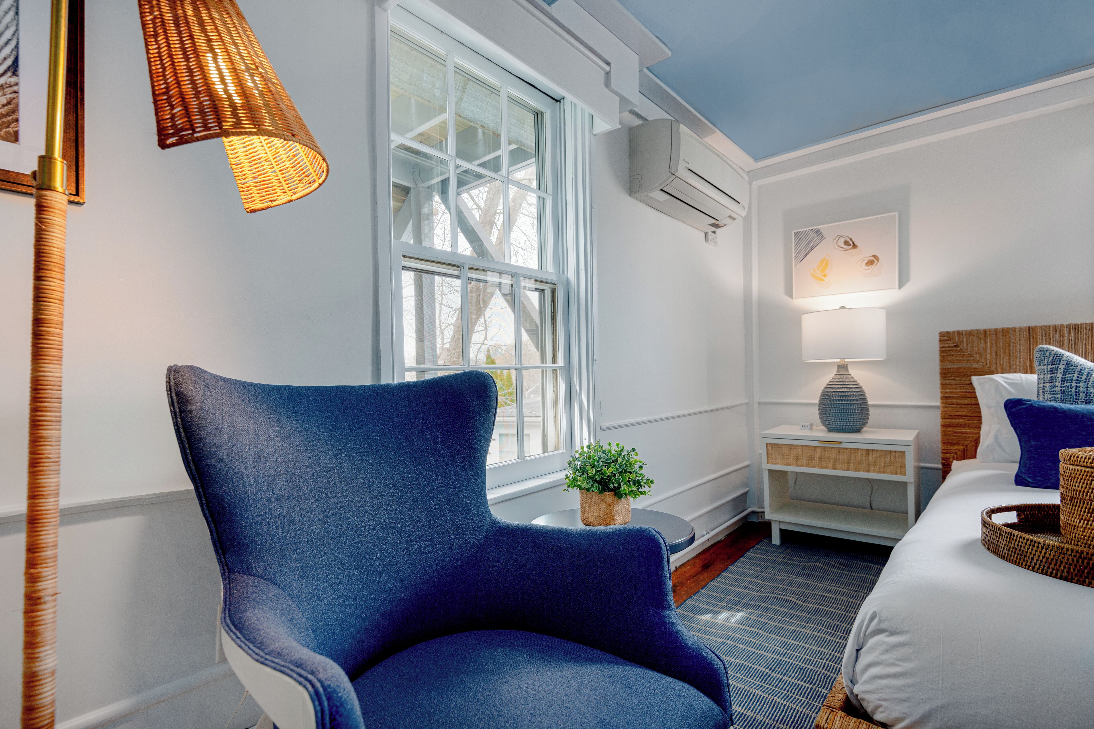 A room with a large window, a blue armchair, a bedside table with a lamp, and a bed with pillows.