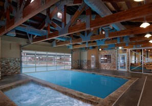 Una piscina interior con borde de baldosas de piedra, vigas de madera y una puerta de cristal que da al exterior.