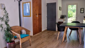 A room with a wooden floor, a wicker chair with a blue cushion, a dining table with chairs, a door, and a potted plant. - Cottage 2/3 pers., Near the sea and sea view, 23 km from Ajaccio (Calcatoggio)