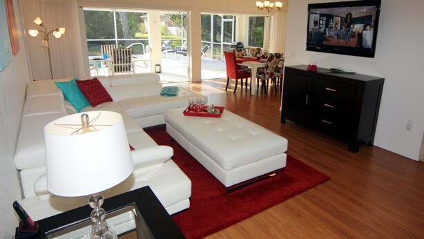 A modern living room with a white sofa, red and blue cushions, a white coffee table, and a red rug. There is a TV mounted on the wall, a ceiling fan, and a dining area with chairs and a table. - Lauras Paradise (Rotonda West)