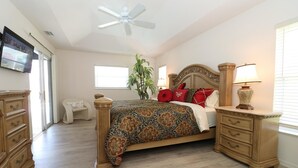 A bedroom with a large bed, a ceiling fan, a nightstand with a lamp, a dresser, and a plant.
