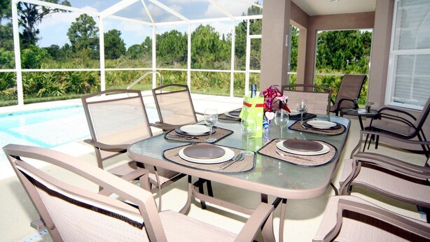 An outdoor dining area with a glass table and chairs, surrounded by a covered patio with a pool view. - Dolphin Cove (Port Charlotte)