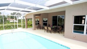 A covered swimming pool area with a patio table and chairs, large windows, and a view of greenery. - Dolphin Cove (Port Charlotte)