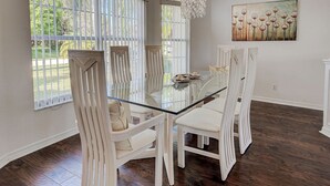 A dining area with a glass-top table and white upholstered chairs, large windows with vertical blinds, and a painting on the wall.
