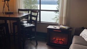 A cozy room with a wooden table, chairs, a fireplace, a sofa, and a large window overlooking a lake.