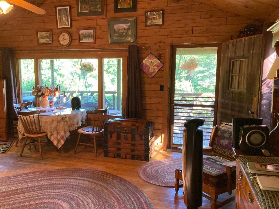 A wooden cabin interior with a dining area, a fireplace, and a large window overlooking greenery.