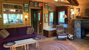 Interior acogedor de una cabaña con piso de madera, un sofá, una silla, una chimenea y un ventilador de techo.