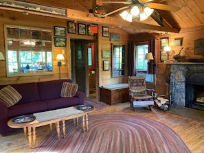 L’intérieur d’un chalet accueillant, avec un plancher en bois, un canapé, un fauteuil, une cheminée et un ventilateur de plafond.