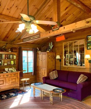 A cozy cabin interior with a wooden ceiling, a ceiling fan, a purple sofa, a wooden table, and a bookshelf.