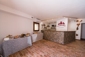 A room with a brick floor, a buffet table with various dishes, a stone counter with a sign reading "Le Undici Rose," and a wooden door. - Le Undici Rose Hotel (Viterbo)