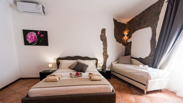 A bedroom with a large bed, a smaller bed, a nightstand with a lamp, a wall-mounted air conditioner, a painting of a rose, and a stone wall feature. - Le Undici Rose Hotel (Viterbo)