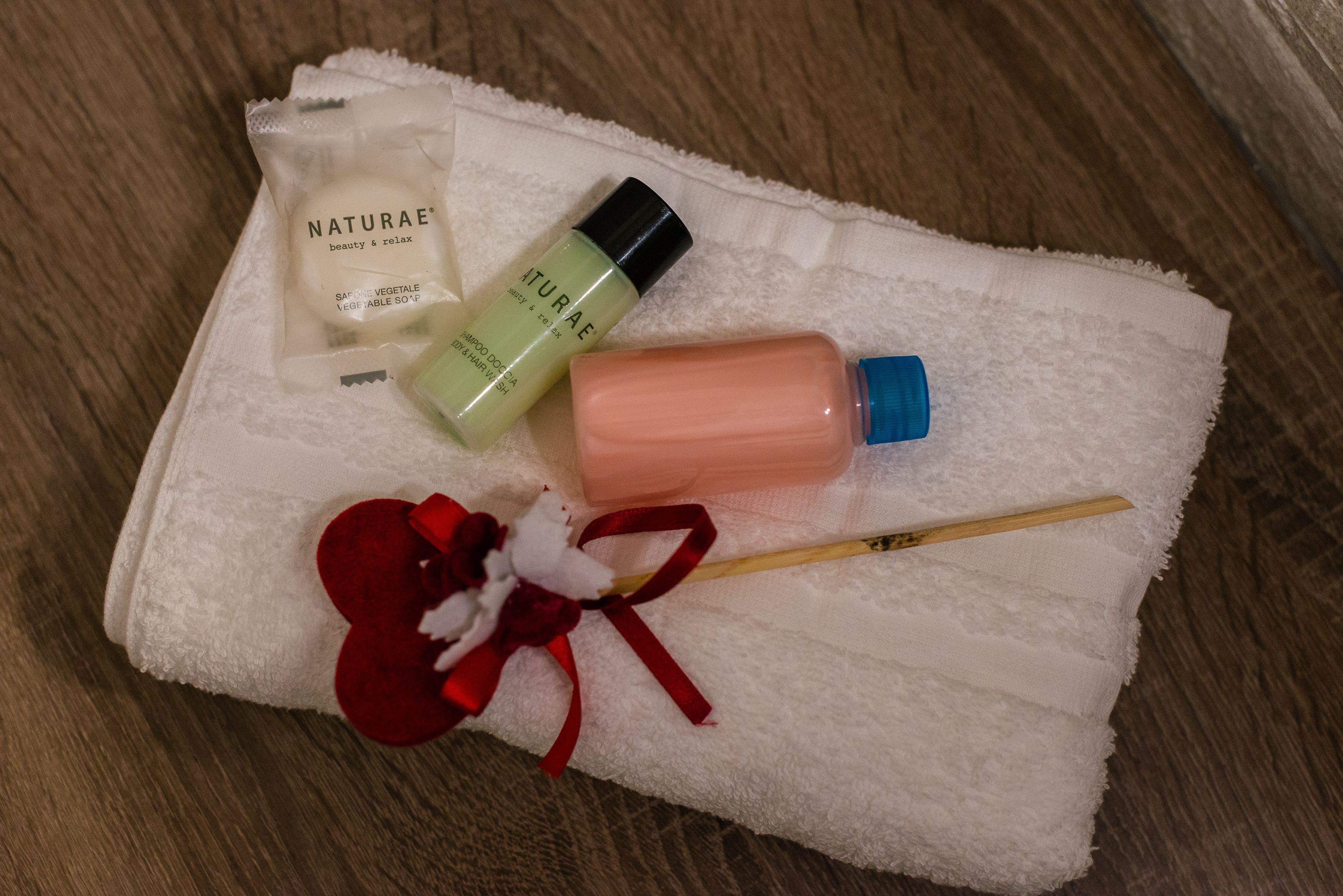 Un asciugamano bianco con un fiore rosso e un bastoncino di legno, insieme a una saponetta di Naturae beauty and relax, una bottiglia di olio vegetale Naturae e una bottiglia rosa.