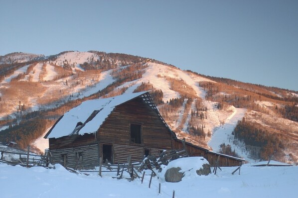 Una cabaña de madera con techo inclinado, rodeada de nieve y una montaña boscosa al fondo.