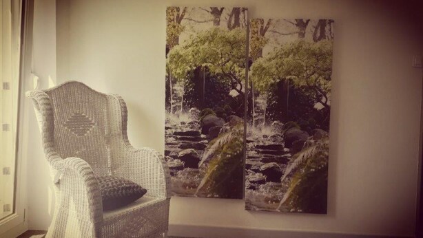 Een rieten stoel in een kamer met een groot schilderij van een waterval en bomen aan de muur.