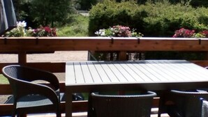 Een houten terras met een tafel en stoelen, omgeven door weelderig groen en bloeiende planten.