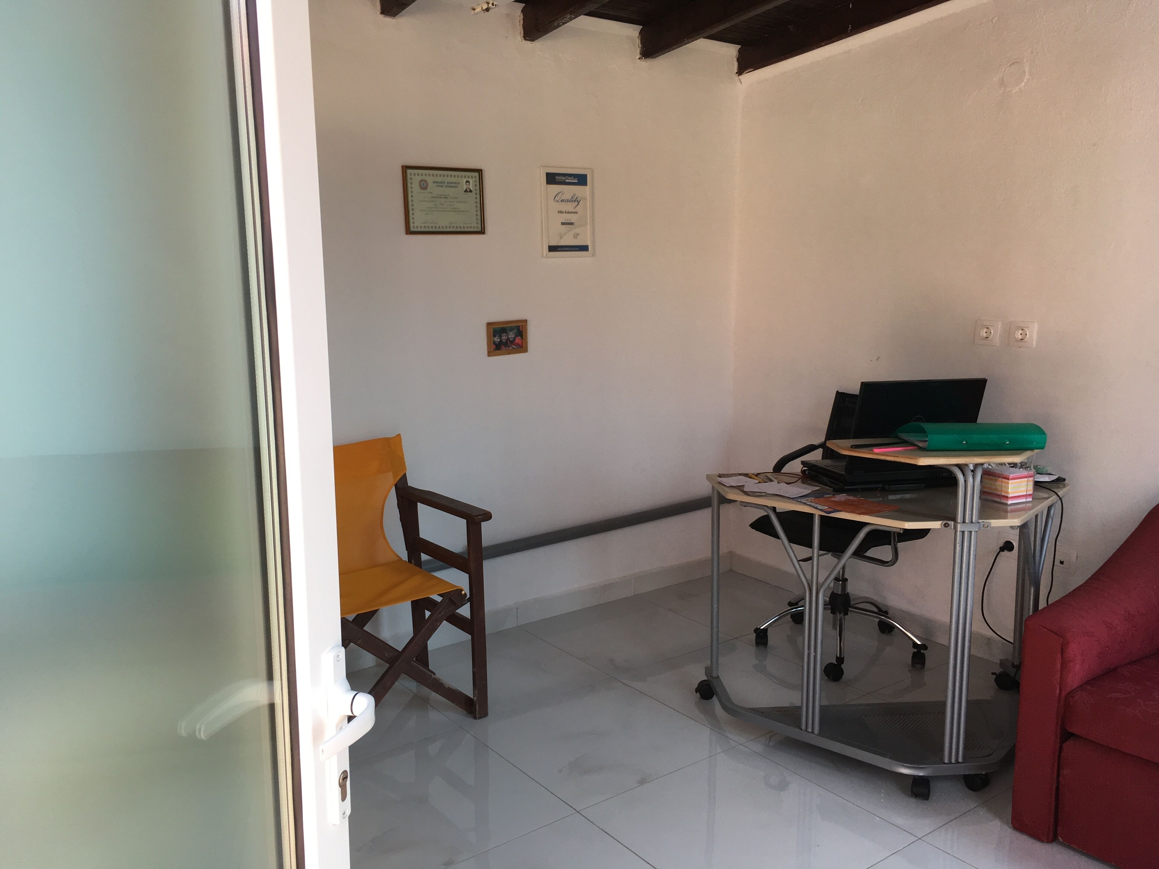 Een kamer met een bureau, stoel en een rode bank. Er hangen certificaten aan de muur en een laptop staat op het bureau.