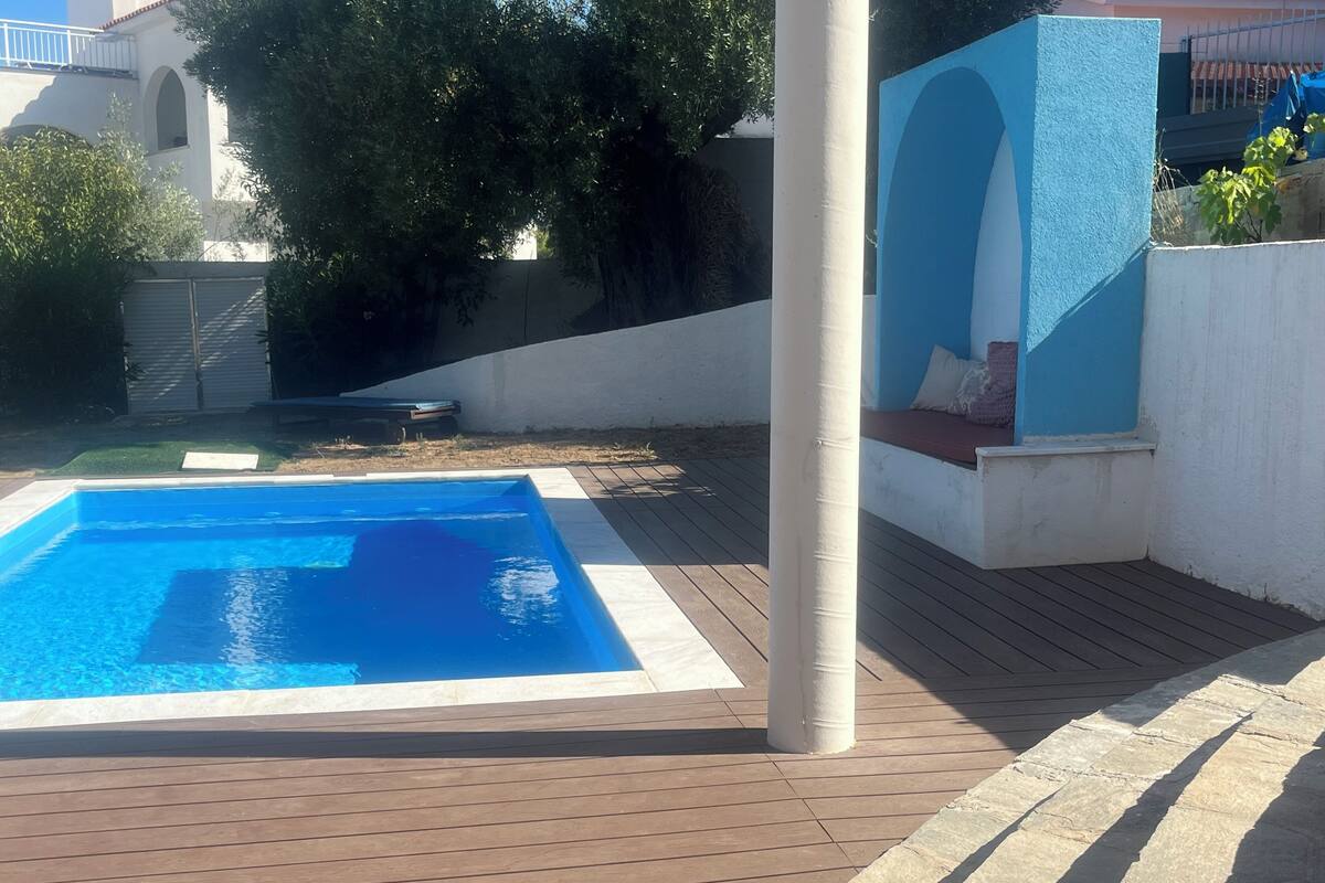 Une piscine avec une terrasse en bois, un pilier blanc et un mur bleu agrémenté d’une fenêtre.