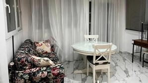 Una habitación con un sofá floral, una mesa redonda de vidrio y sillas, y un ventanal con cortinas.