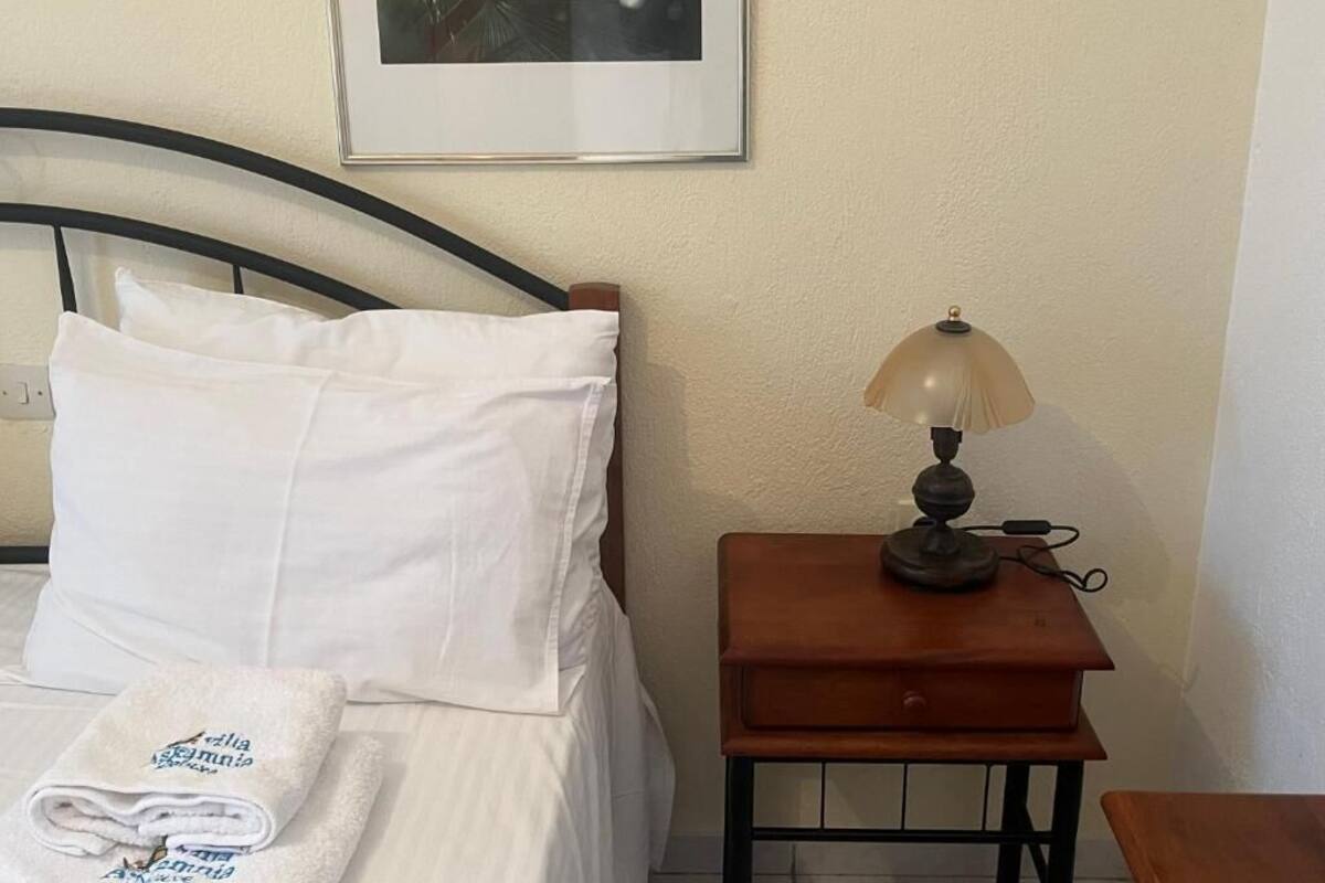 Un lit bien fait, avec du linge de lit blanc et une serviette pliée ornée d’un motif, une table de chevet avec une lampe et un tableau encadré accroché au mur.
