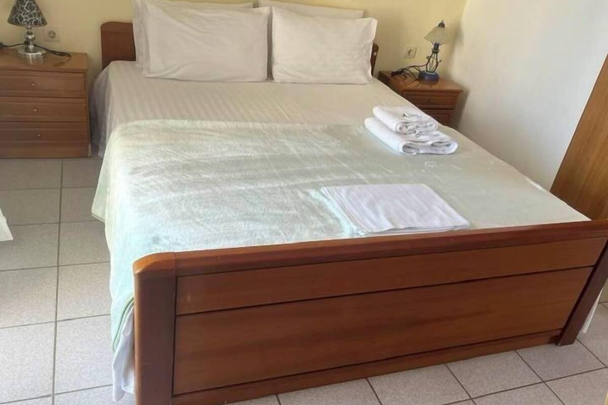 Un lit bien fait, avec un cadre en bois, une couvre-lit blanche et deux oreillers. Il y a deux tables de chevet avec des lampes et une serviette pliée sur le lit.