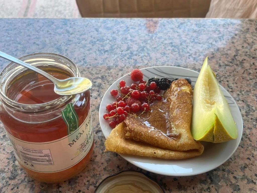 Ein Teller mit Pfannkuchen, Beeren und einem Melonenschnitzchen, sowie ein Marmeladenglas auf einer Granit-Arbeitsplatte.