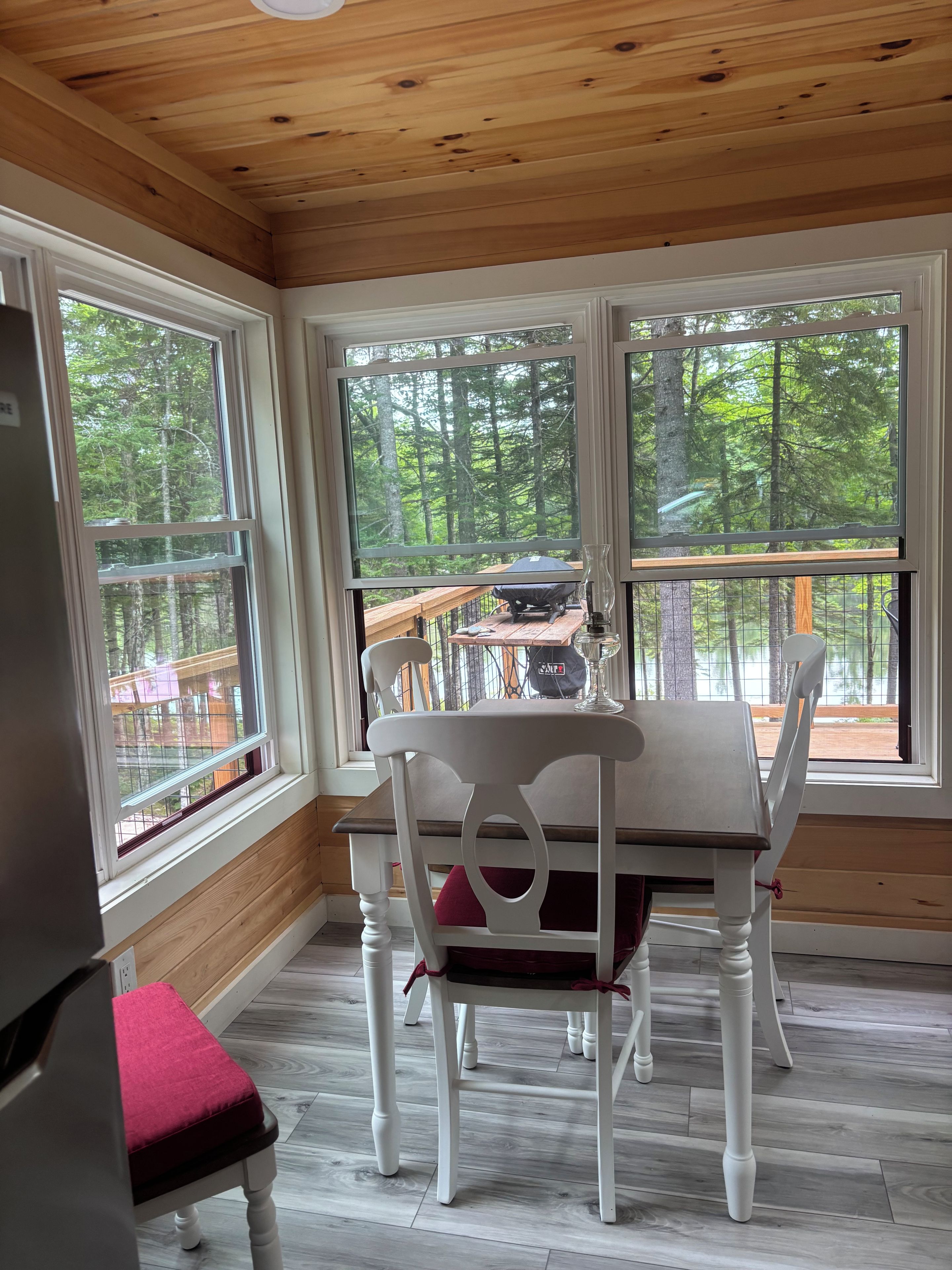 A dining area with a table and chairs, large windows with a view of a forest, and a wooden ceiling.